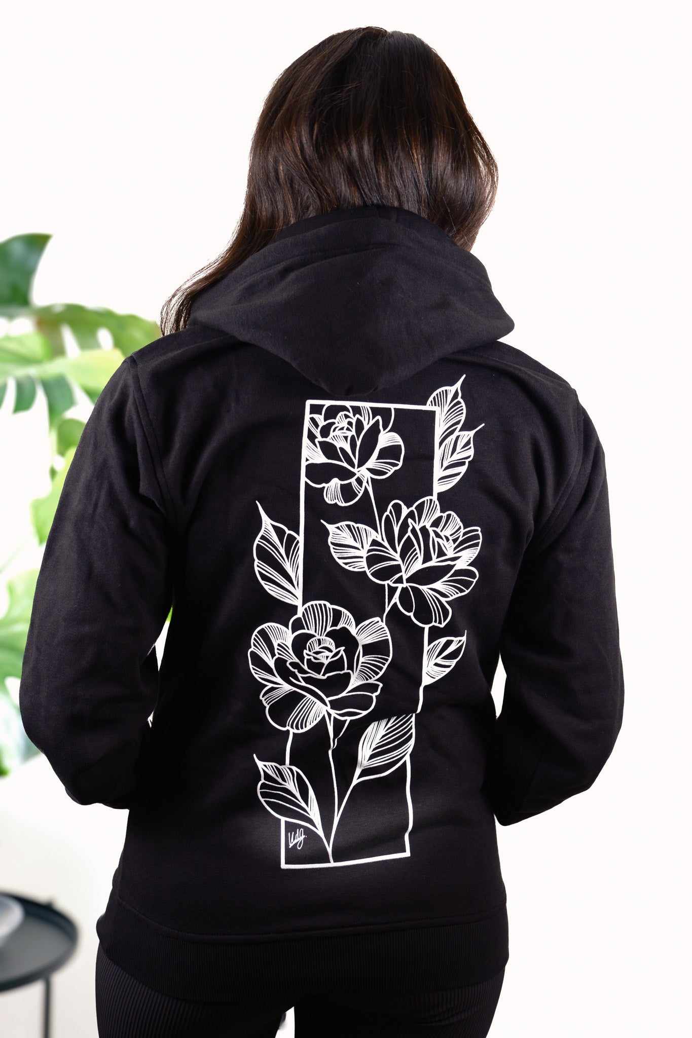 Female model wearing Love Knows No Bounds hoodie, back view featuring heart and floral design inspired by fine line tattoo art.