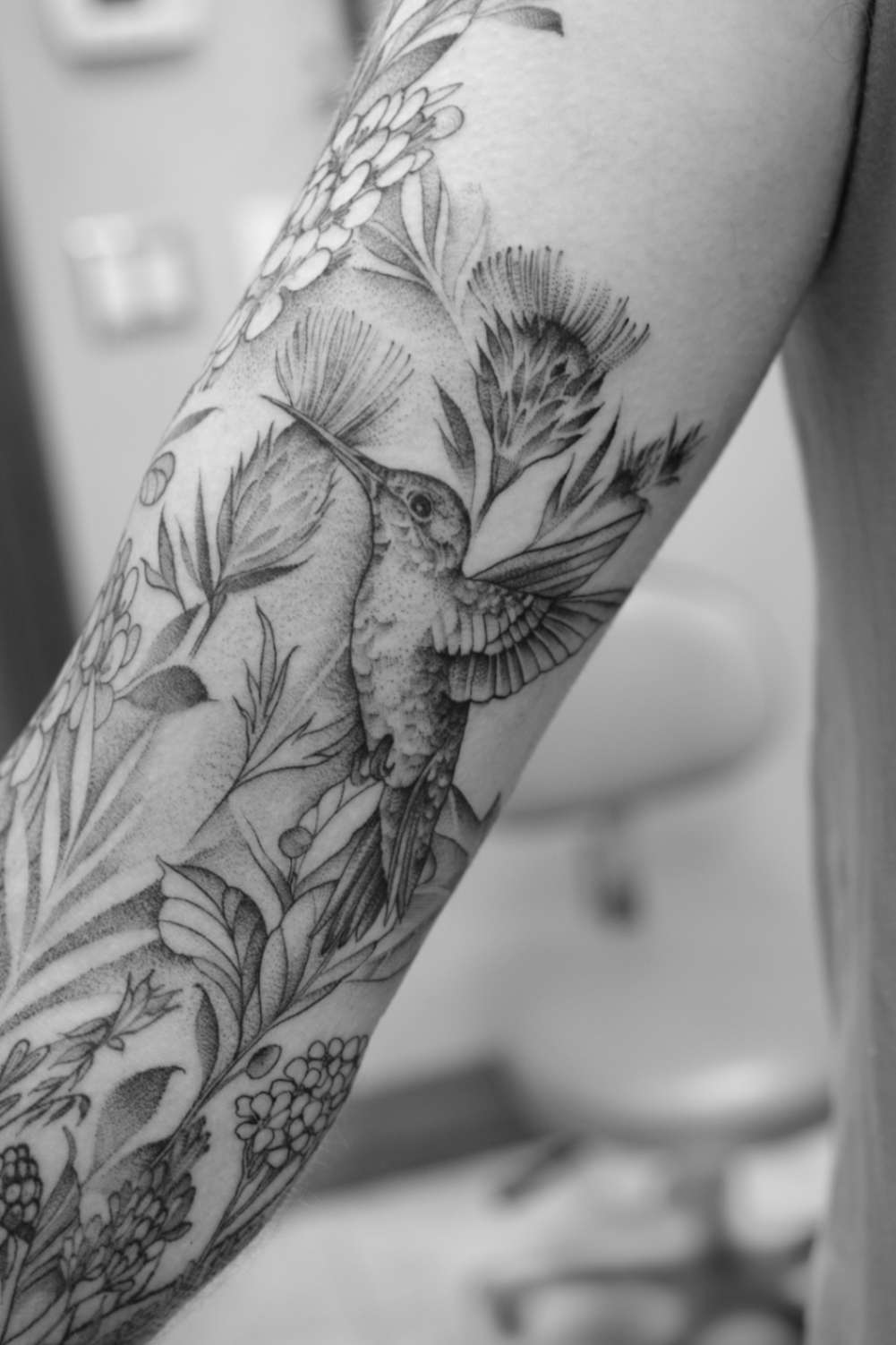 Banner image of a fine line floral and hummingbird tattoo sleeve on a woman's arm, featuring delicate lines and subtle shading, displayed on the tattoo convention page.