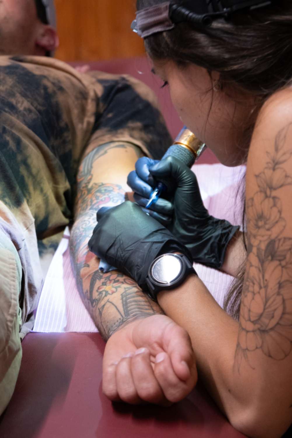 Kansinee meticulously executing fine line tattoo work on a man's right inner arm, demonstrating her precision and skill with a tattoo machine in creating detailed tattoo designs.