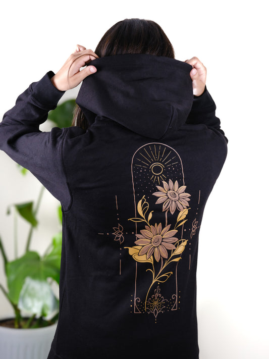 Female model wearing Follow the Sun hoodie, back view featuring vibrant sunflower design with fine line detailing