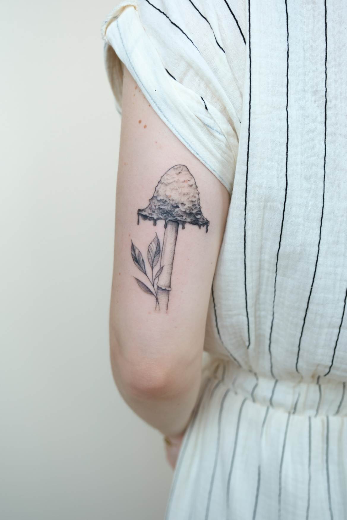 Fine line tattoo of an ink cap mushroom and floral design on a woman's left tricep, highlighting delicate artistic details.